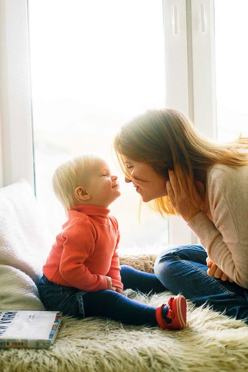 A mother and her young child touching noses while sitting near a window.